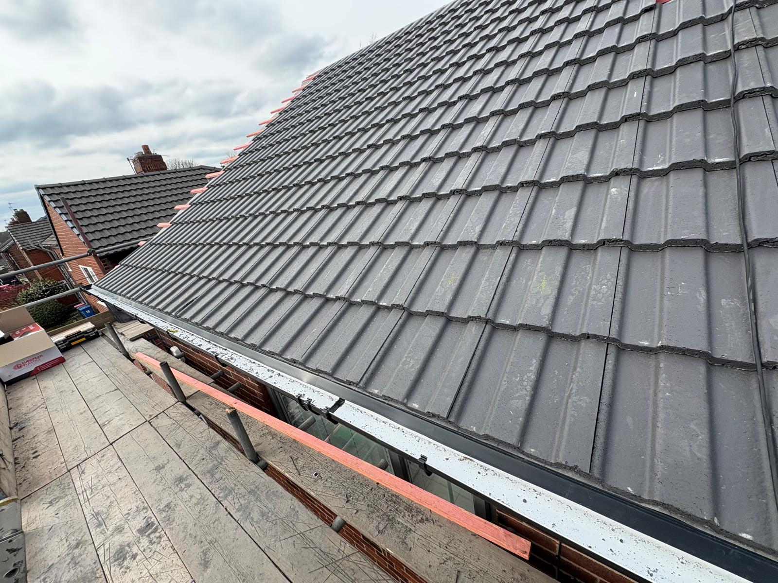 Newly laid grey profiled tiles running down to the eaves, with a new white fascia and black gutter below and scaffold boards in the foreground